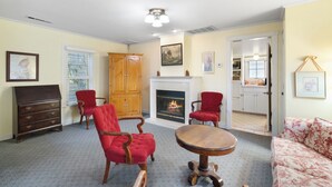 A living room with a fireplace, red upholstered chairs, a wooden table, and a floral sofa. - Cottage 9 – Downtown Jacksonville Retreat Steps from Shops & Britt Gardens (Jacksonville)