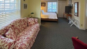 A room with a floral sofa, a bed with white bedding, a small table, and a television. - Cottage 9 – Downtown Jacksonville Retreat Steps from Shops & Britt Gardens (Jacksonville)