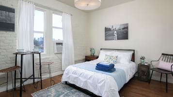 A bedroom with a bed, a table, two chairs, a rug, a nightstand, and a wall-mounted lamp.