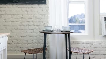 A room with a brick wall, a window with a view, a table with two chairs, and a small shelf with a kettle.