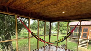 Una terraza de madera con una hamaca, ventanas amplias y vistas a una casa y árboles.