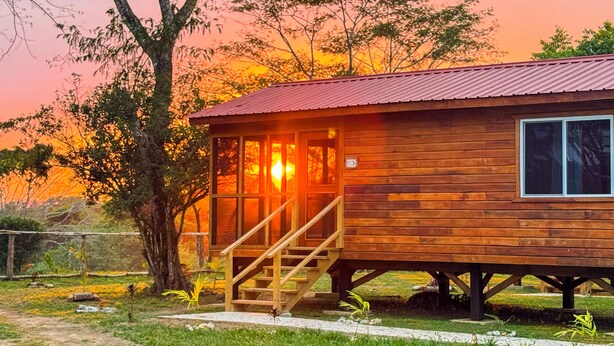 Una cabaña de madera con techo de metal, una escalera que lleva a la entrada y una vista del atardecer al fondo.