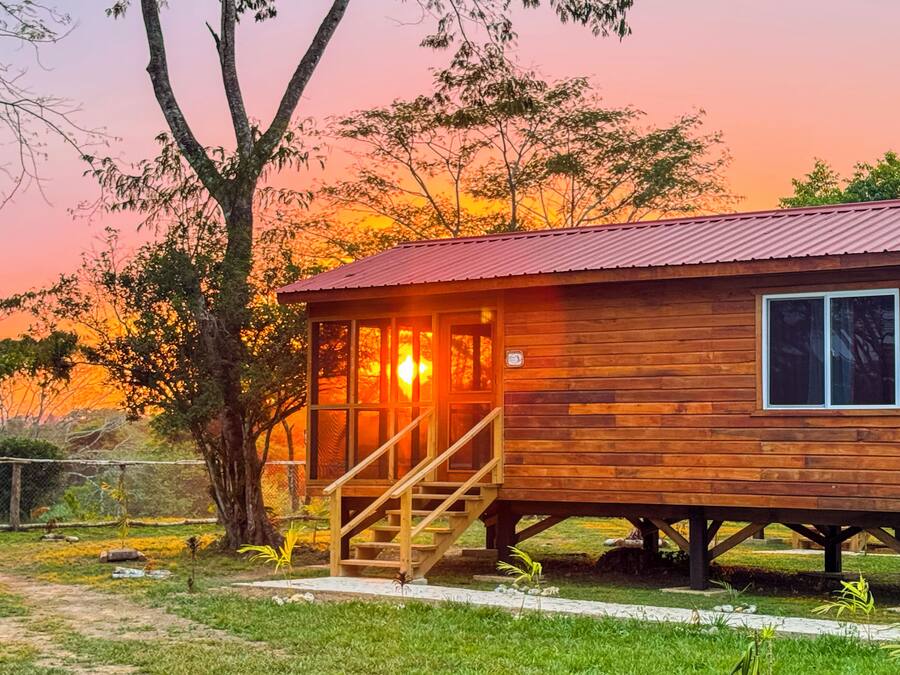 Una cabaña de madera con techo de metal, una escalera que lleva a la entrada y una vista del atardecer al fondo.