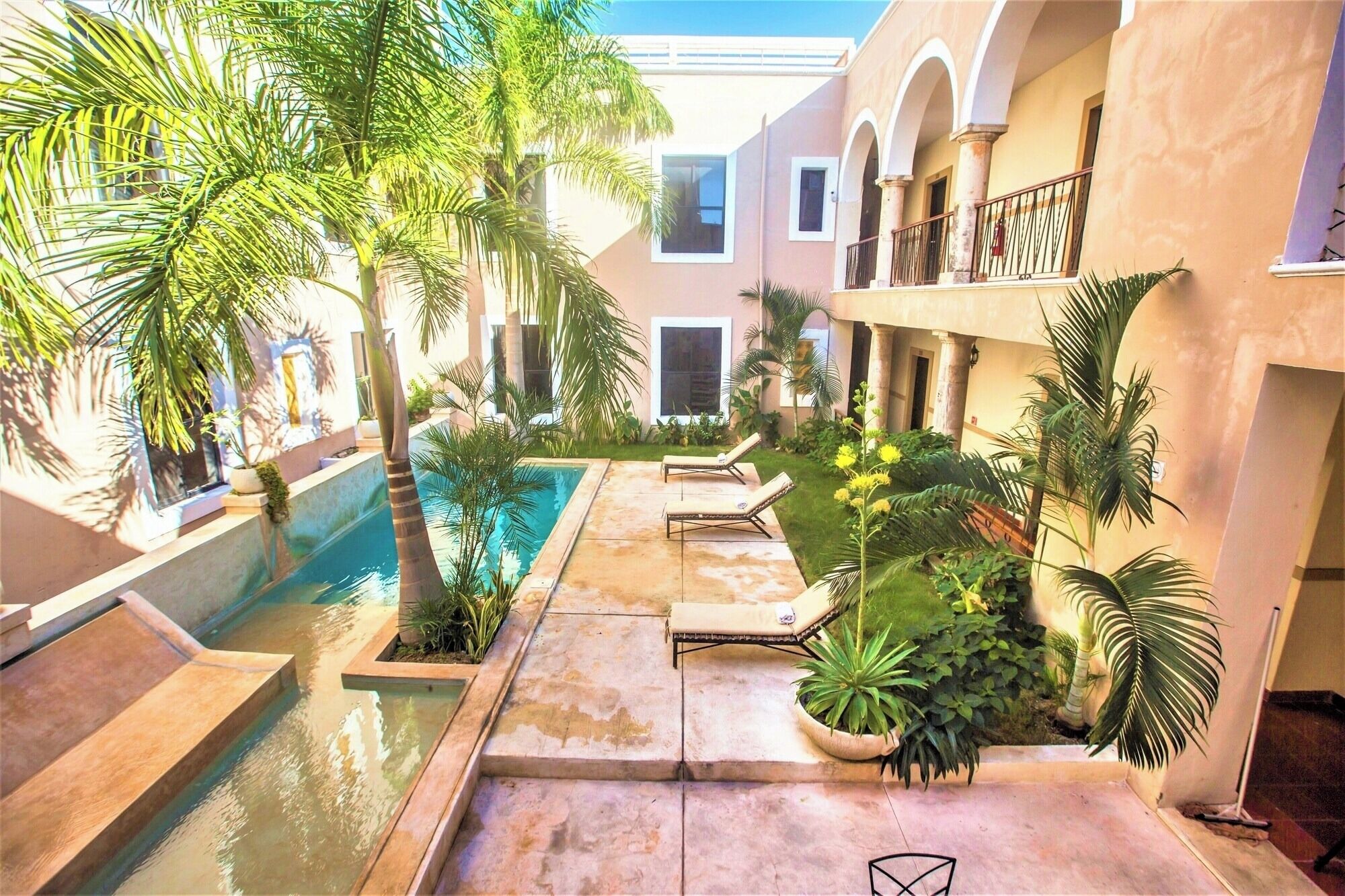 Zona de piscina con sillones, palmeras y un edificio con arcos y balcones.