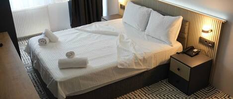 A neatly made bed with white linens and two rolled towels, a bedside table with a phone, and a patterned rug.