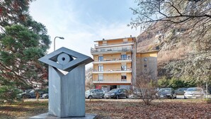 Een modern gebouw met balkon, een parkeerplaats met auto's en een sculptuur op de voorgrond.