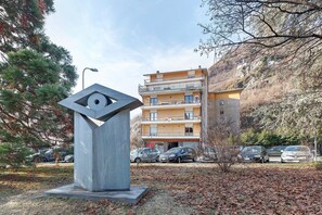 Un edificio moderno con un balcón, un estacionamiento con coches y una escultura al frente.