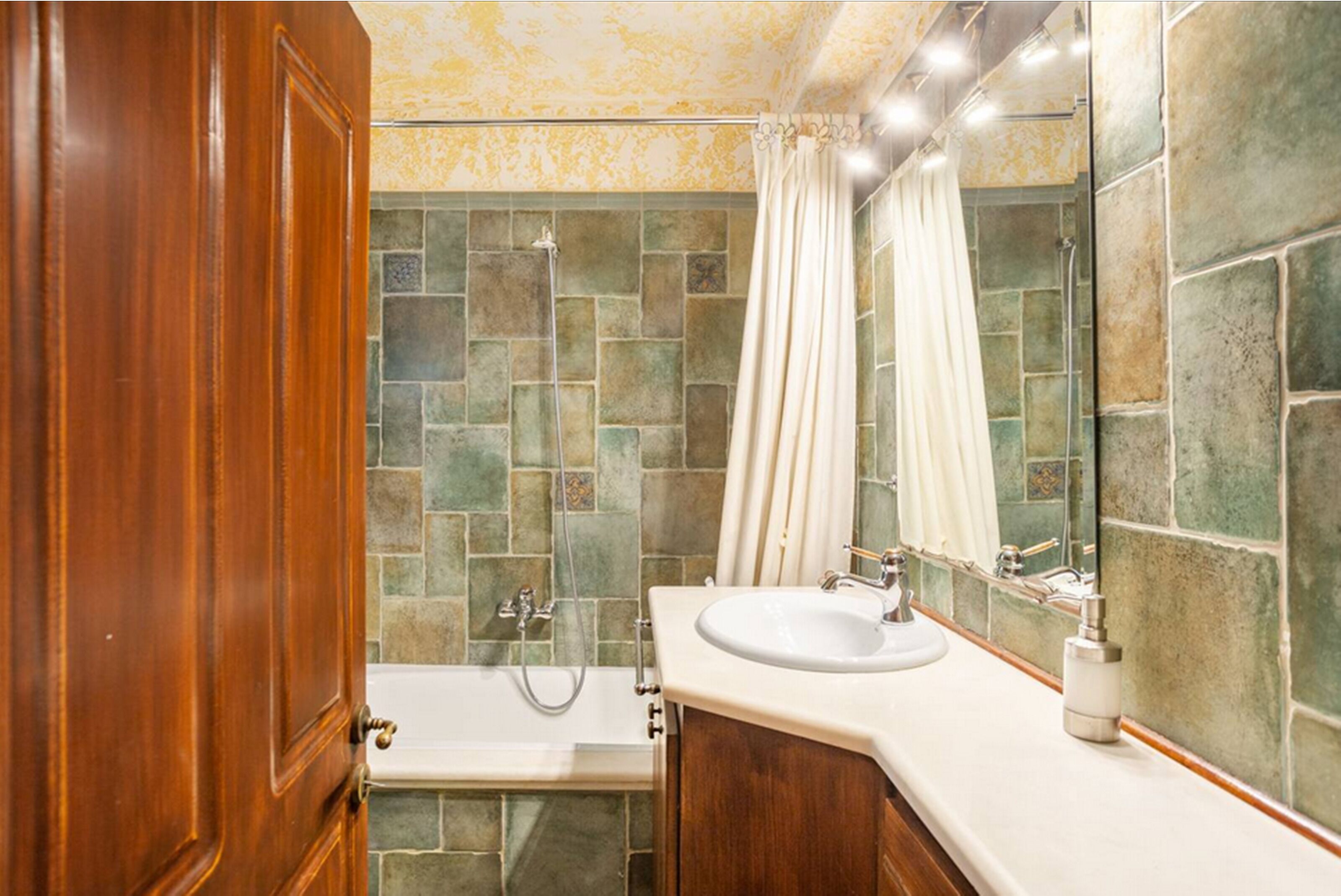 A bathroom with a stone-tiled shower wall, a wooden door, a white sink, and a mirror.