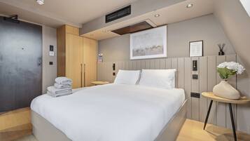 A modern hotel room with a large bed, a wooden wardrobe, a small round table with a vase, and a framed picture on the wall.