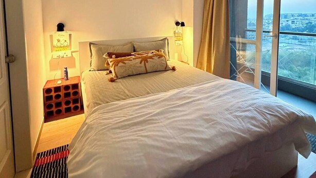 Camera d'albergo con un letto grande, una ventola a soffitto, un comodino con una lampada e vista sulla città attraverso ampie finestre.