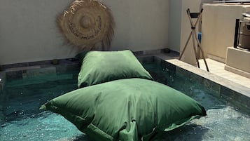 Une piscine sur le toit avec une eau cristalline, deux coussins verts, un chapeau de paille et une petite structure blanche.