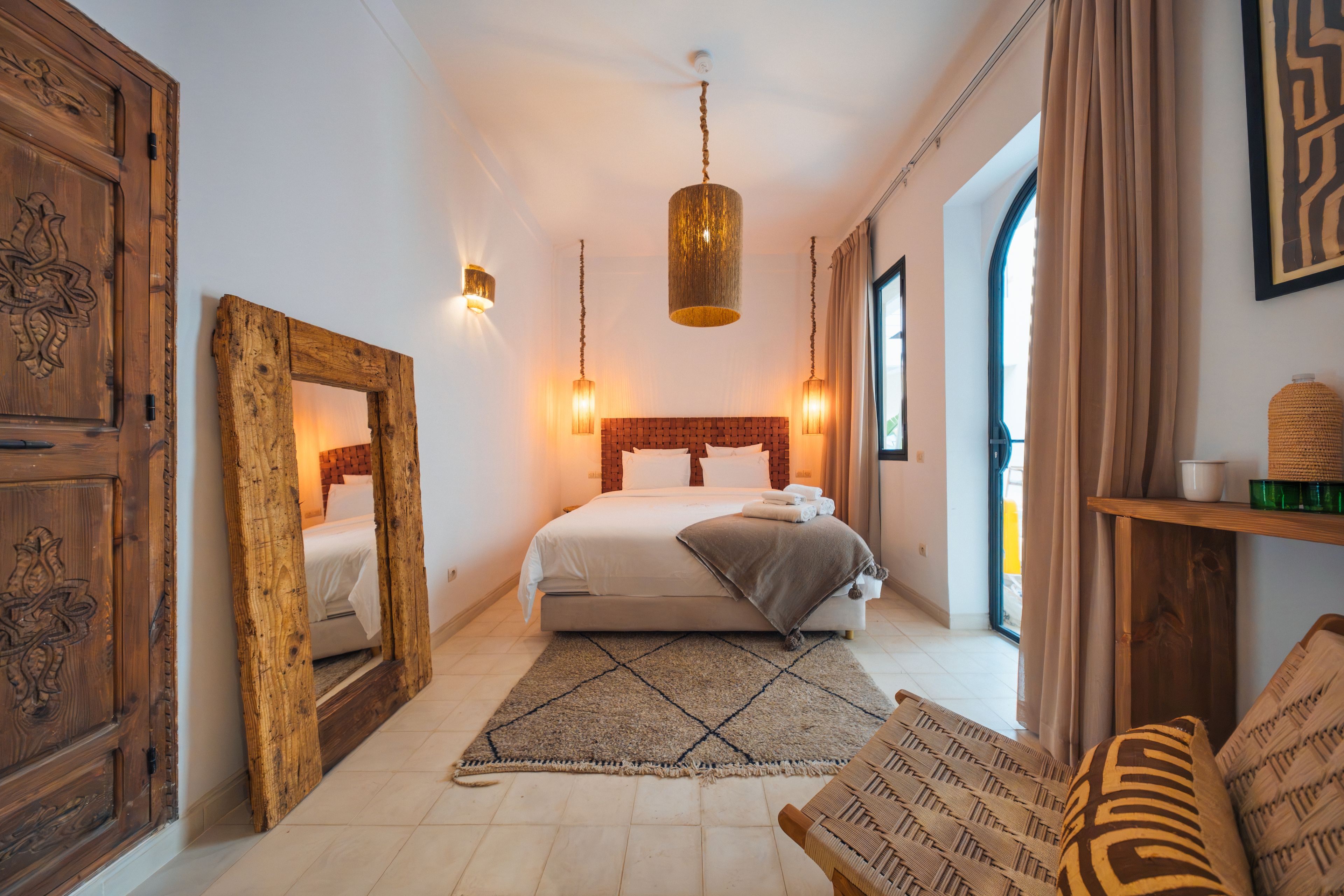 A hotel room with a large bed, a wooden wardrobe, a hanging light fixture, and a view of the outside.