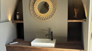 A bathroom with a white sink, a black faucet, a round mirror with a geometric design, and wooden shelves. - Riad A l'Ombre du Palais (Marrakech)