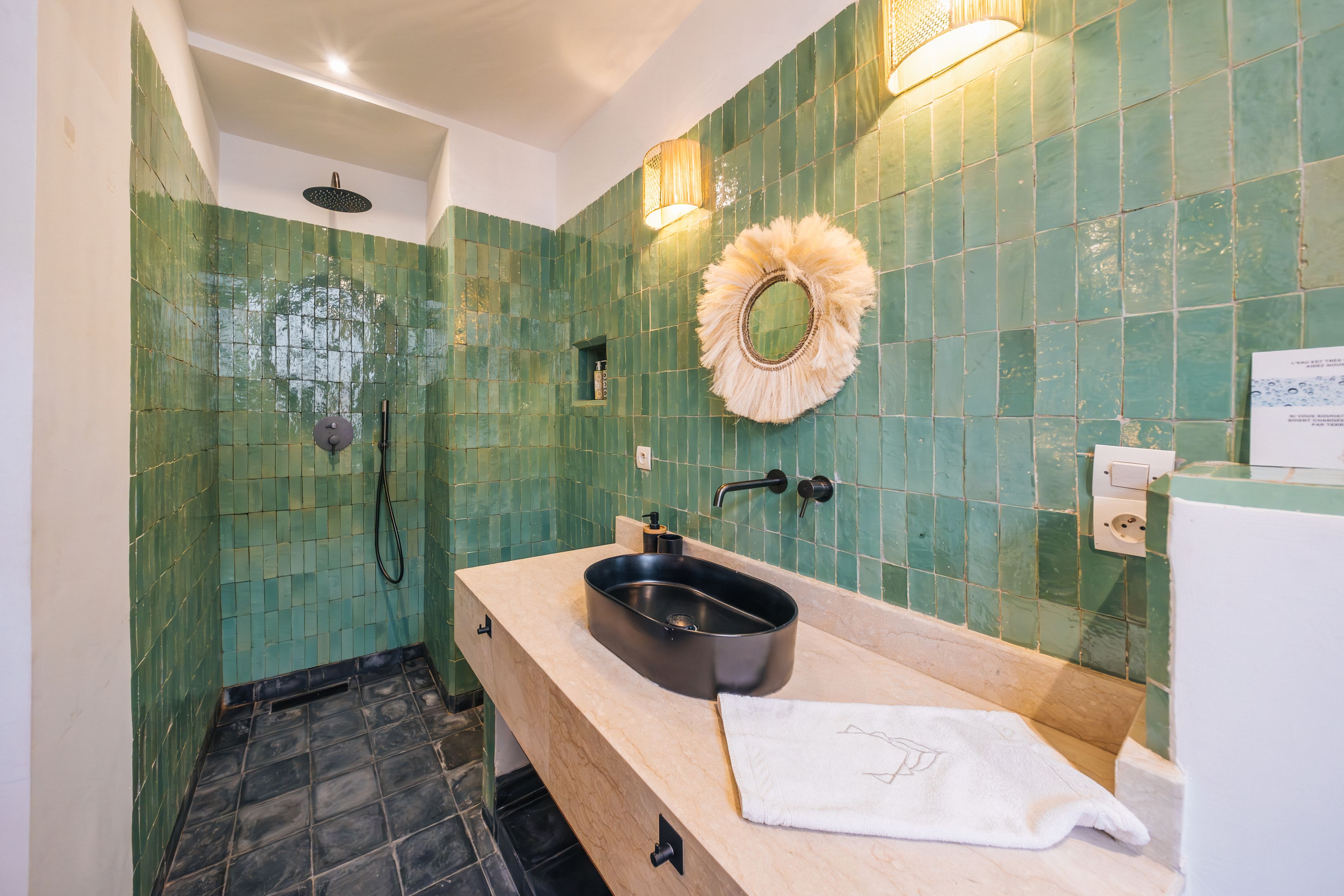 Casa de banho com parede em azulejos verdes, lavatório preto, toalha e chuveiro.