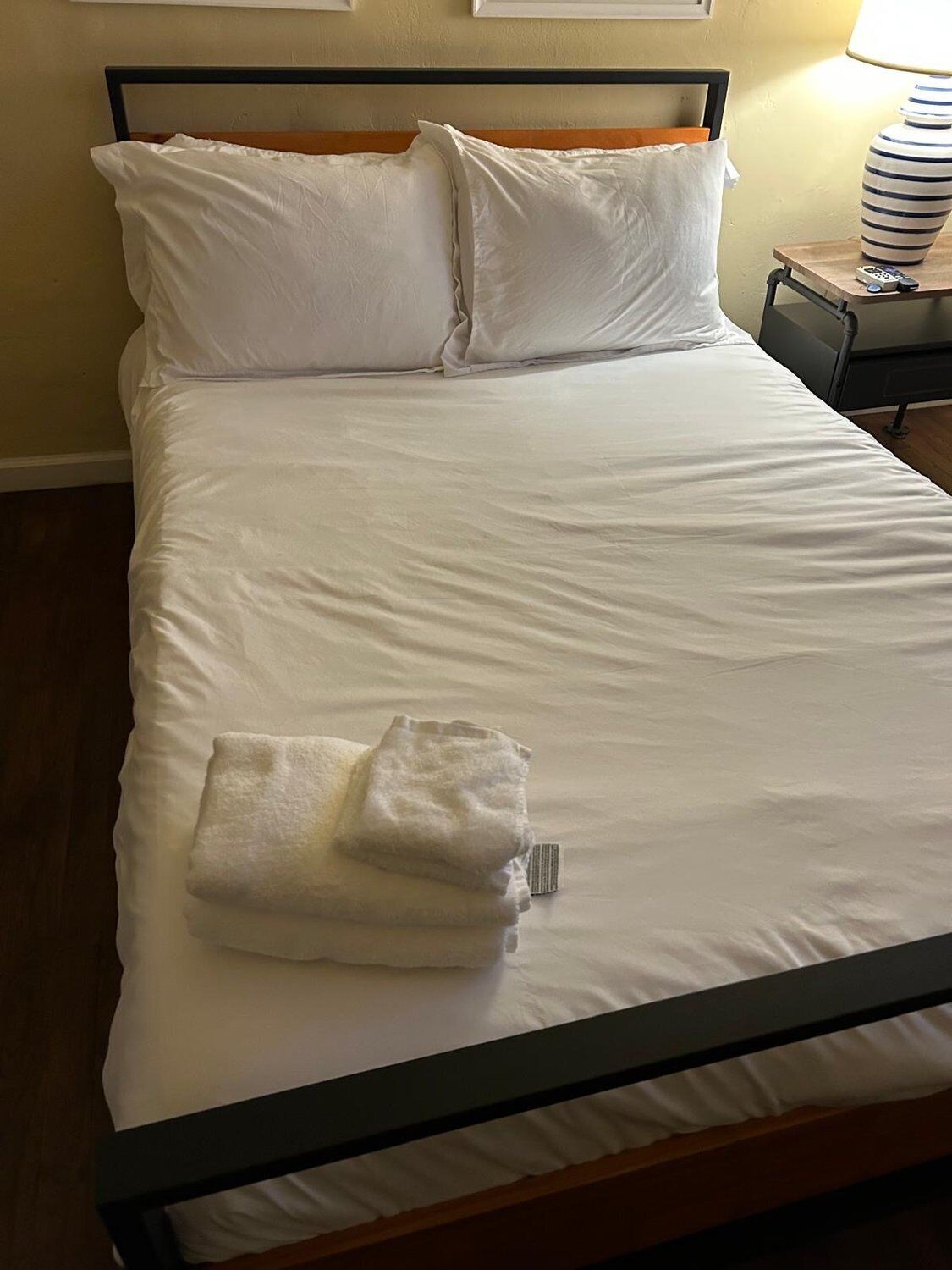 A neatly made bed with white linens and pillows, a folded towel on the bed, and a bedside table with a lamp and remote control.