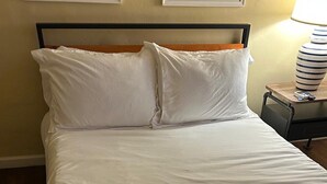 Een net opgemaakt bed met witte lakens en kussens, een opgevouwen handdoek op het bed, en een nachtkastje met een lamp en afstandsbediening.