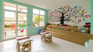 A classroom with a large alphabet tree mural, a play area visible through the glass doors, and a wooden bench.
