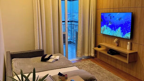 A hotel room with a bed, a TV, a small table, a plant, and a balcony with curtains.