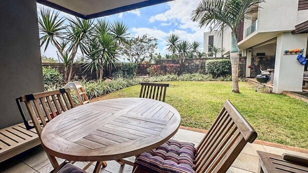 Une terrasse avec une table ronde en bois et des chaises, entourée d’un jardin luxuriant agrémenté de palmiers, avec une maison visible au loin.