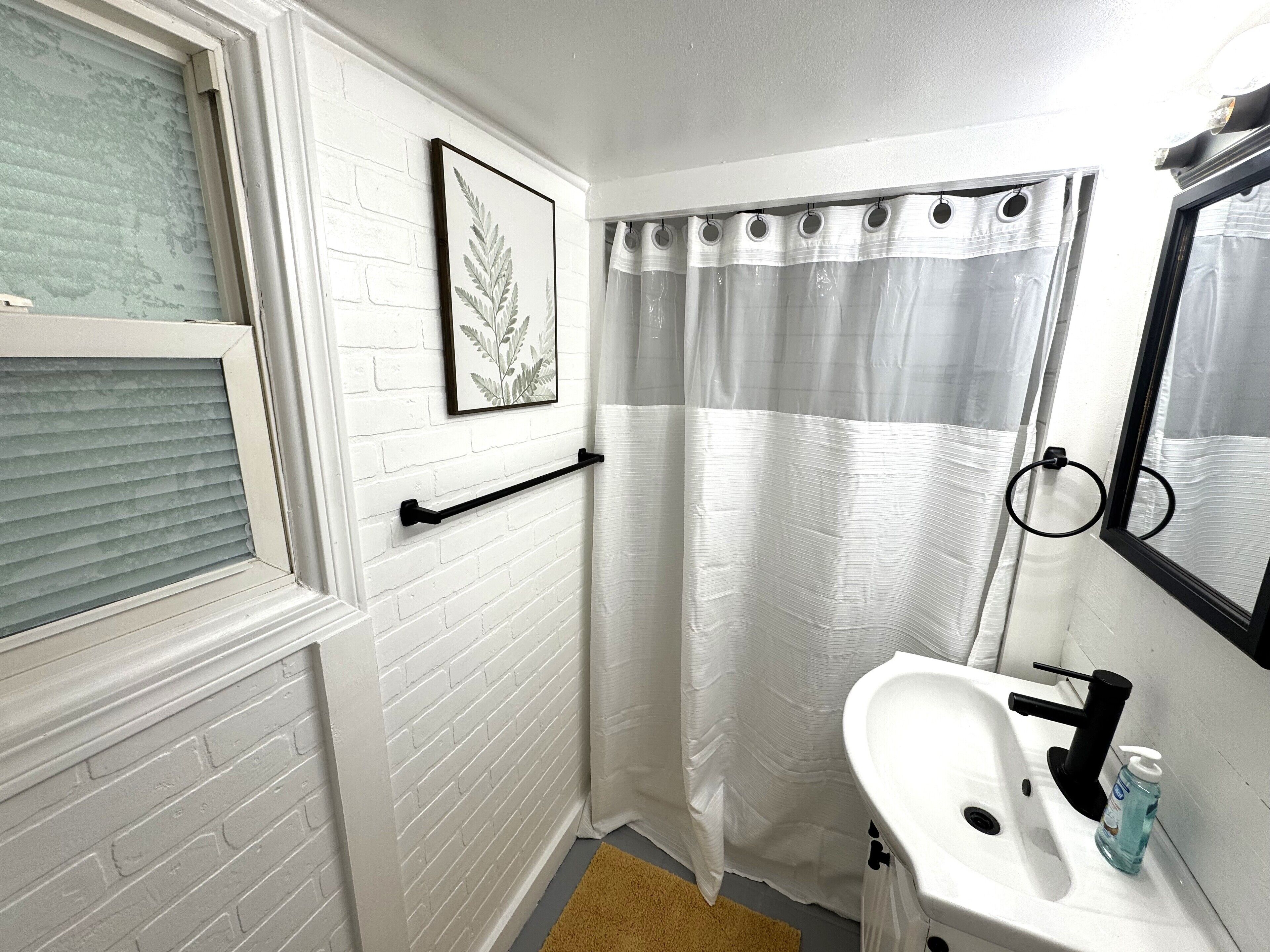 Ein Badezimmer mit weißer Ziegelwand, einem Fenster mit Jalousien, einem Duschvorhang, einem Waschbecken mit Spiegel und einem Handtuchhalter.