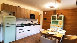 A compact kitchen with a refrigerator, sink, and wooden cabinets. There is a dining table with chairs and a small cabinet with glass doors. - Chalet Peter Serrada, a few meters from the slopes, in villa surrounded by forest (Serrada)