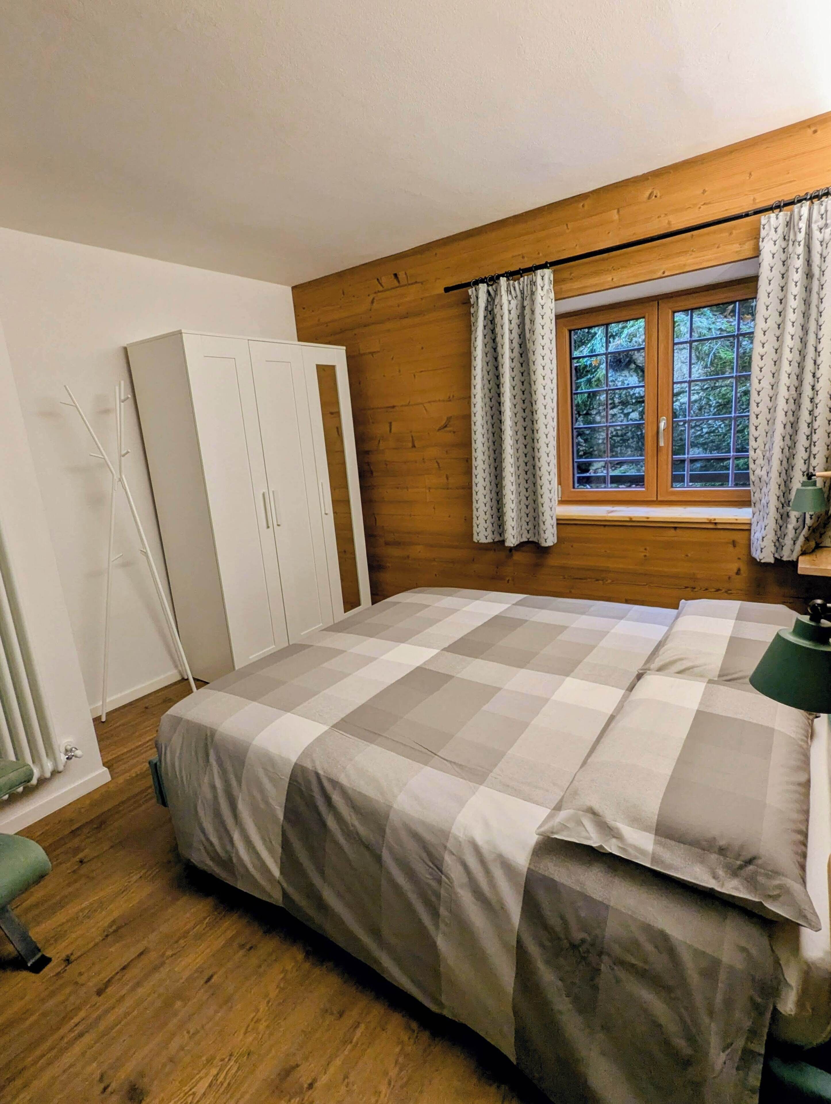 A bedroom with a wooden ceiling, a large bed with a checkered bedspread, a wardrobe, and a window with curtains.