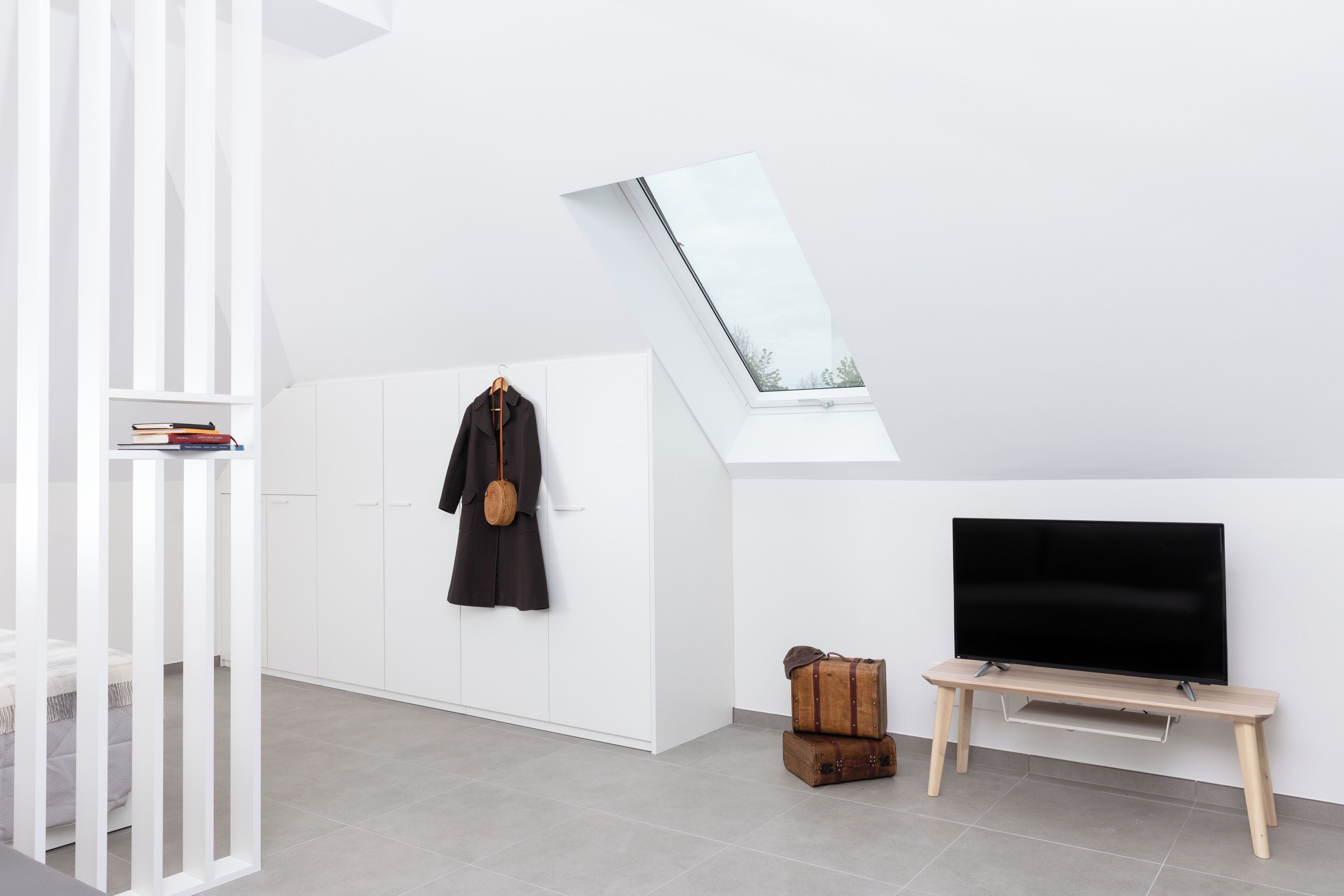 A modern living room with a white wardrobe, a coat hanging on the wall, a television on a wooden stand, and a skylight above.