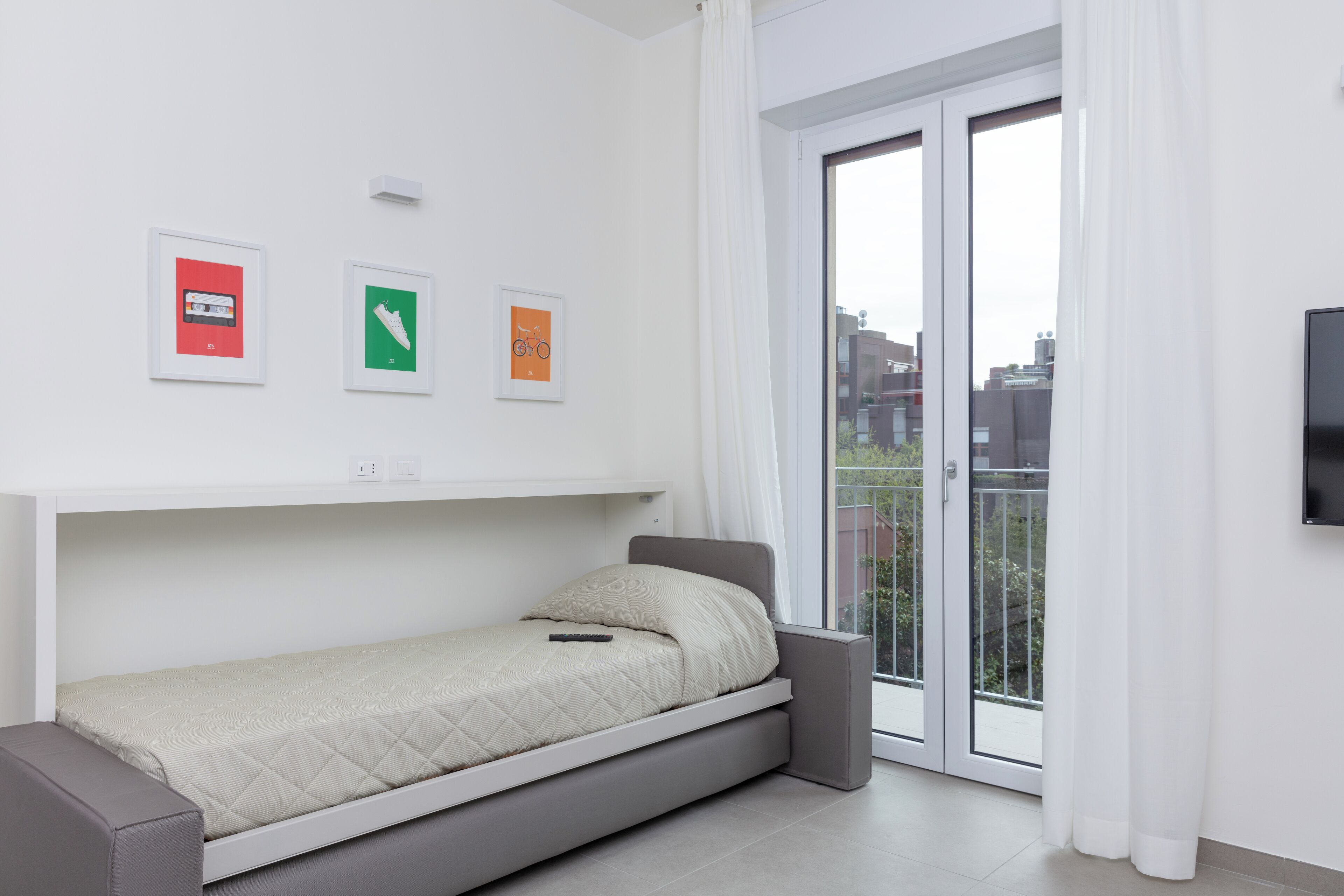 A modern bedroom with a single bed, a sliding door to a balcony, and three framed pictures on the wall.