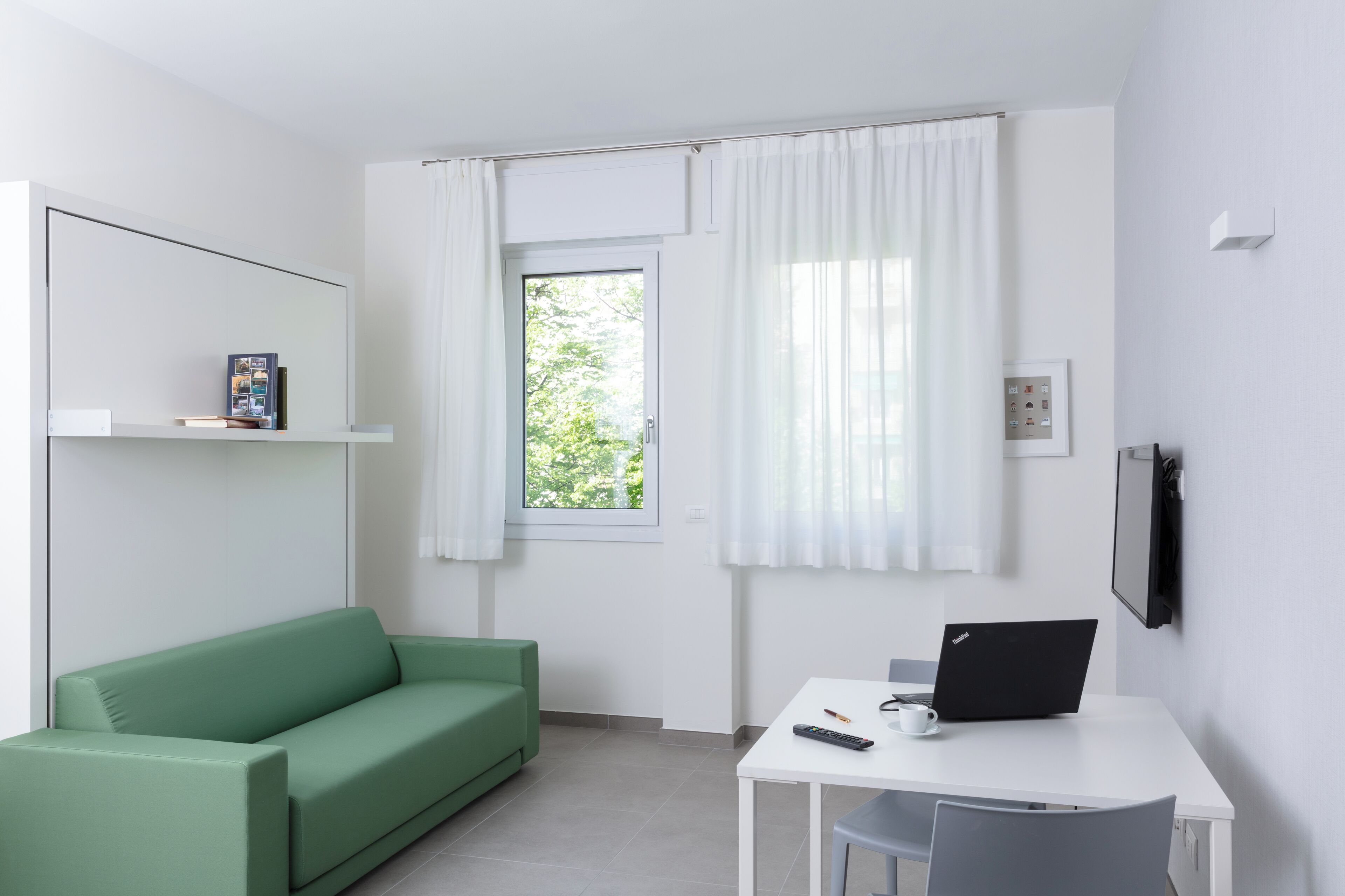 Une pièce moderne avec un canapé vert, un bureau blanc sur lequel se trouve un ordinateur portable, une télécommande et une fenêtre ornée de rideaux blancs.