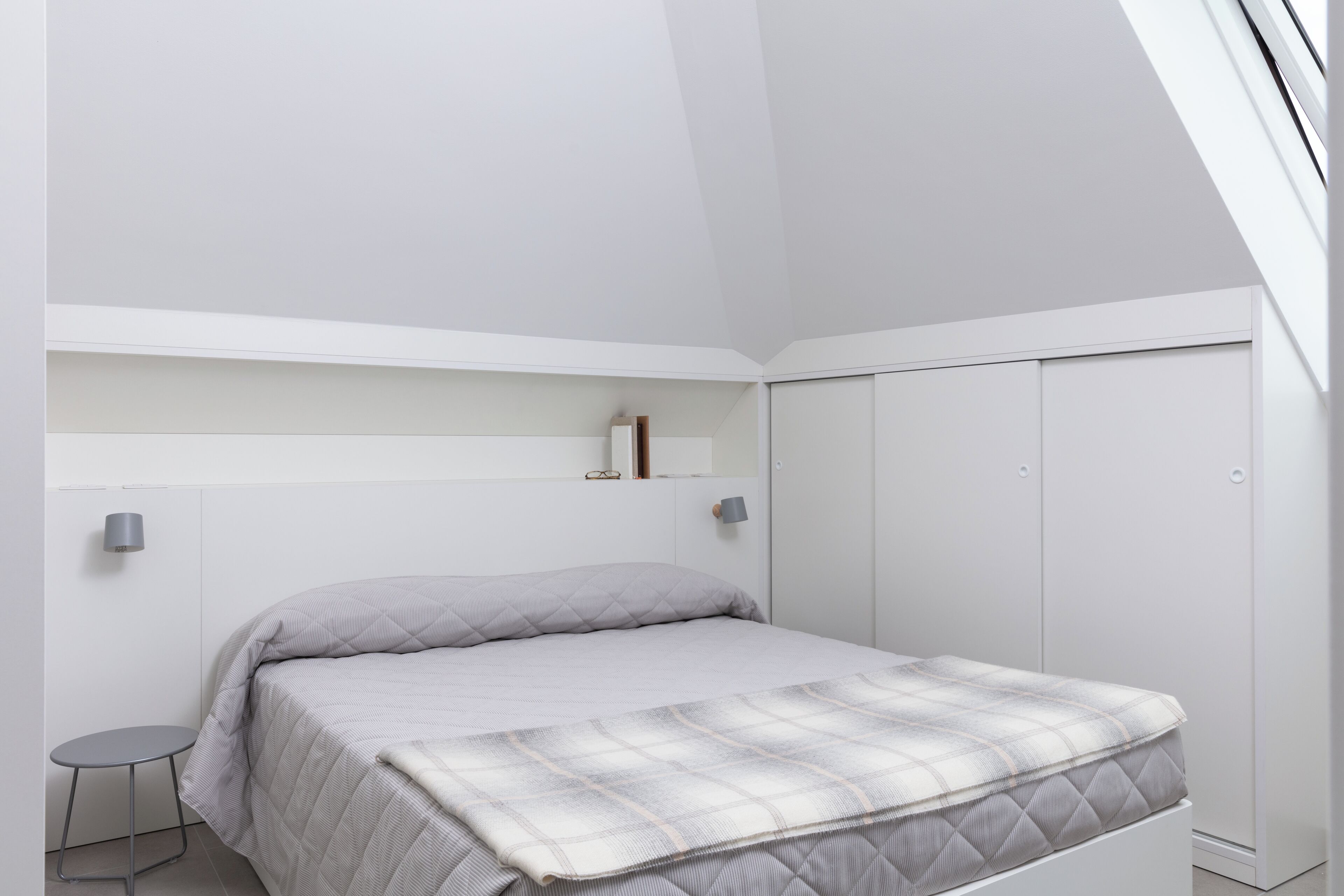 A bedroom with a bed, a small table, a wardrobe, and a shelf with a lamp.