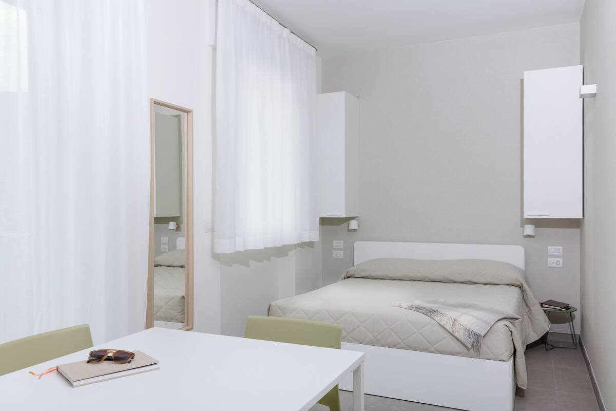 A minimalist bedroom with a bed, a bedside table, a chair, a mirror, and a window with curtains.
