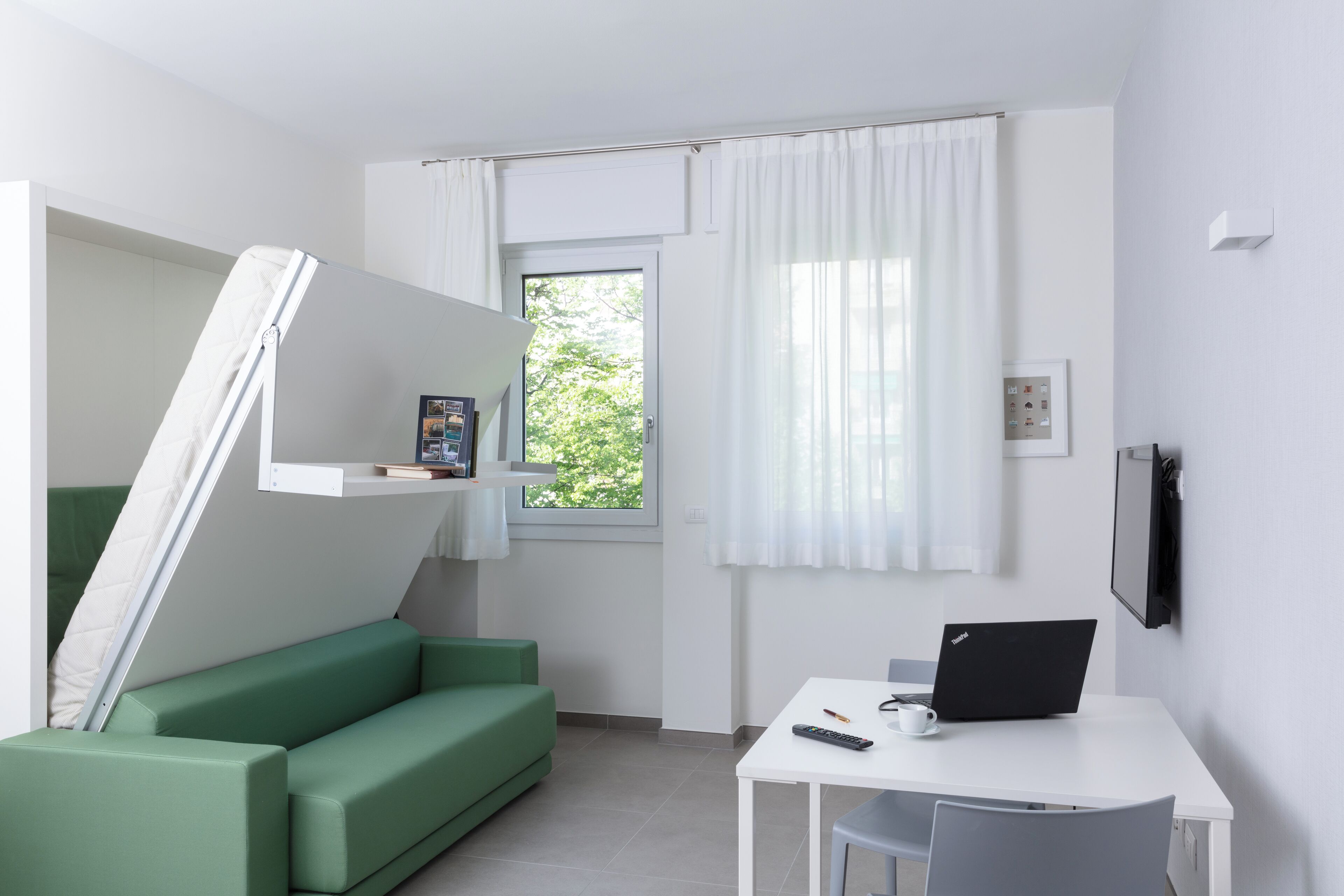 Ein kleines Zimmer mit einem grünen Sofa, einem weißen Schreibtisch, einem Laptop, einer Fernbedienung und einem kleinen Bilderrahmen.