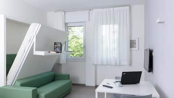 Una habitación compacta con un sofá verde, un escritorio blanco con una computadora portátil, un control remoto y un pequeño marco de fotos.