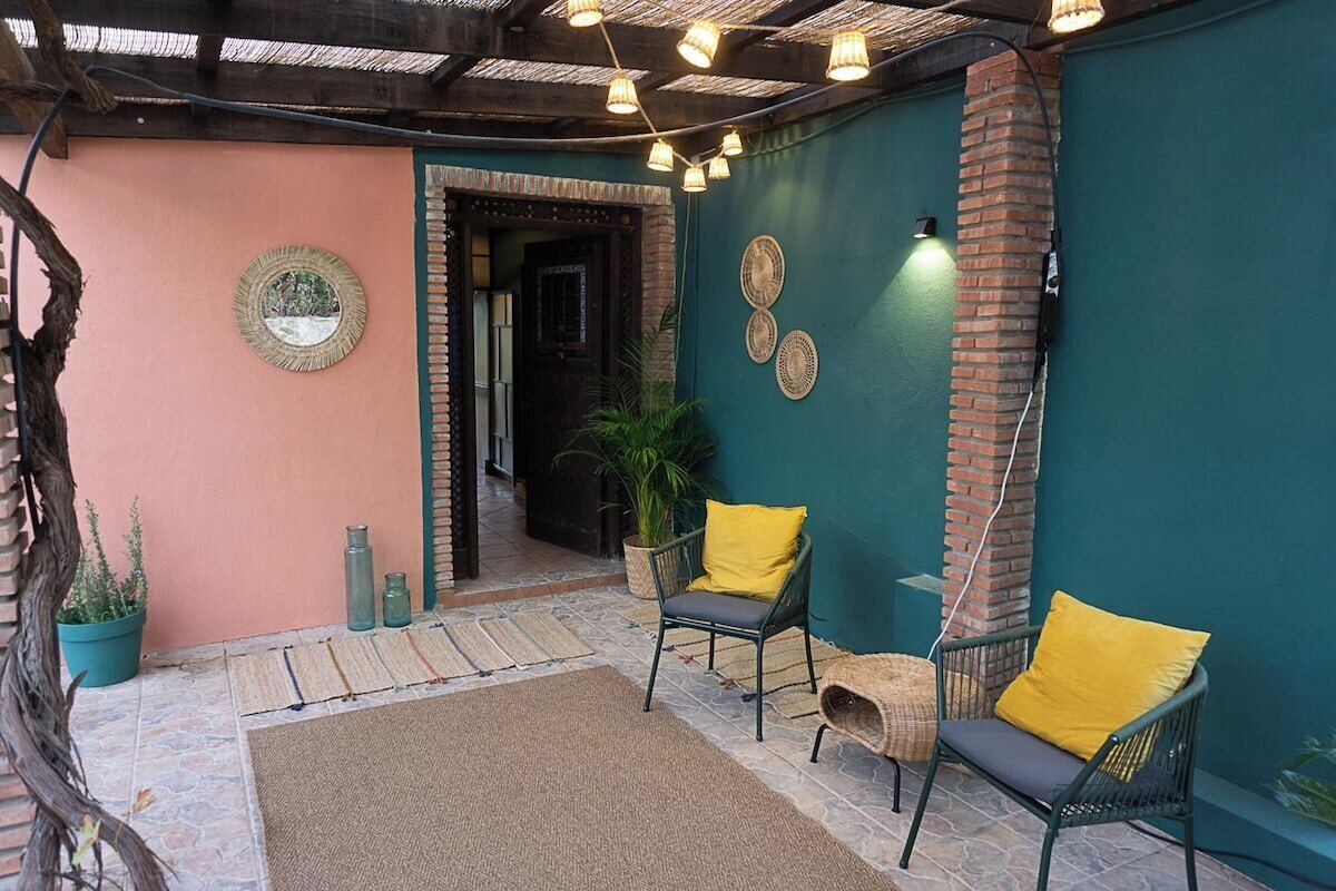 Un espacio al aire libre acogedor con sillas de mimbre, un tapete tejido, un espejo decorativo y una pared de ladrillo.