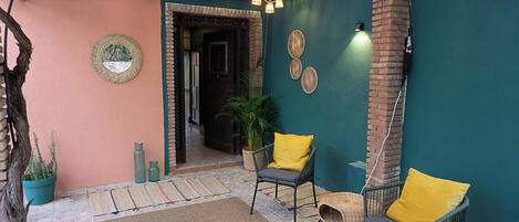 Un espacio al aire libre acogedor con sillas de mimbre, un tapete tejido, un espejo decorativo y una pared de ladrillo.