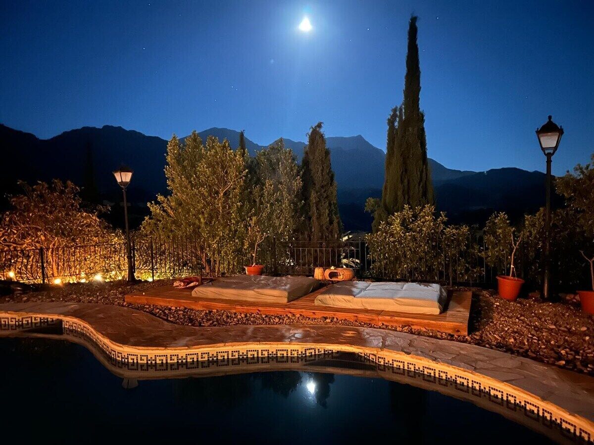 Un espace piscine nocturne, illuminé par la lune, entouré d’arbres et de plantes en pot, avec une chaîne de montagnes en arrière-plan.