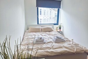 Un lit bien fait, agrémenté d’oreillers et de serviettes, une fenêtre donnant sur des immeubles et une petite table sur laquelle est posée une plante.