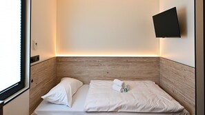 Un petit lit bien fait, avec du linge de lit blanc et des oreillers, un téléviseur à écran plat fixé au mur et une fenêtre avec des stores.