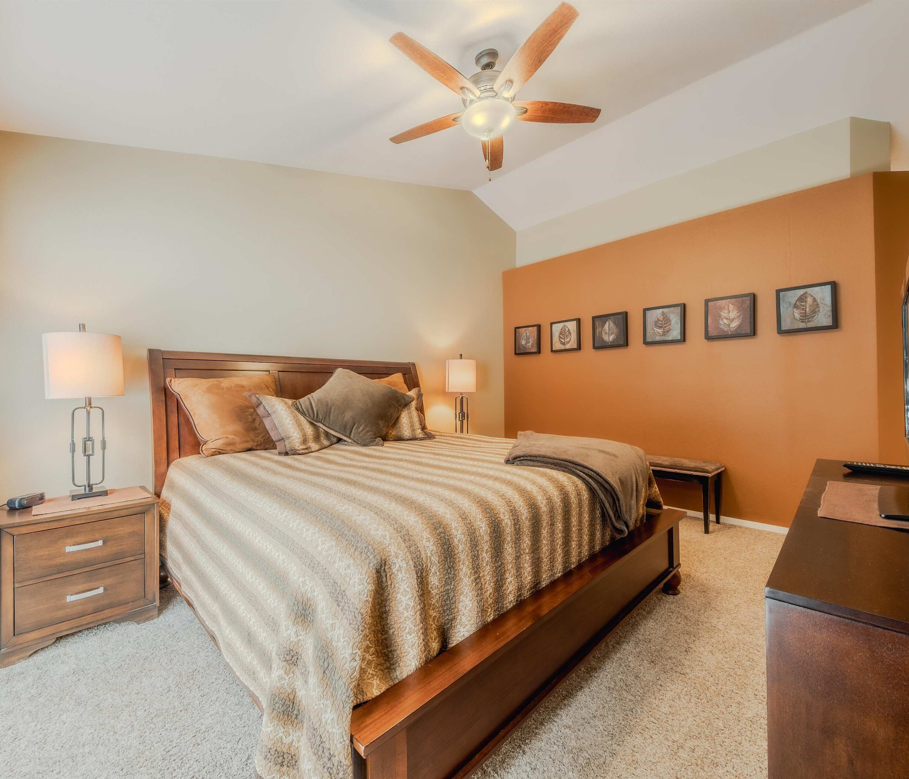 Un dormitorio con una cama de madera, dos mesitas de noche con lámparas, un ventilador de techo y una pared decorada con obras de arte enmarcadas.
