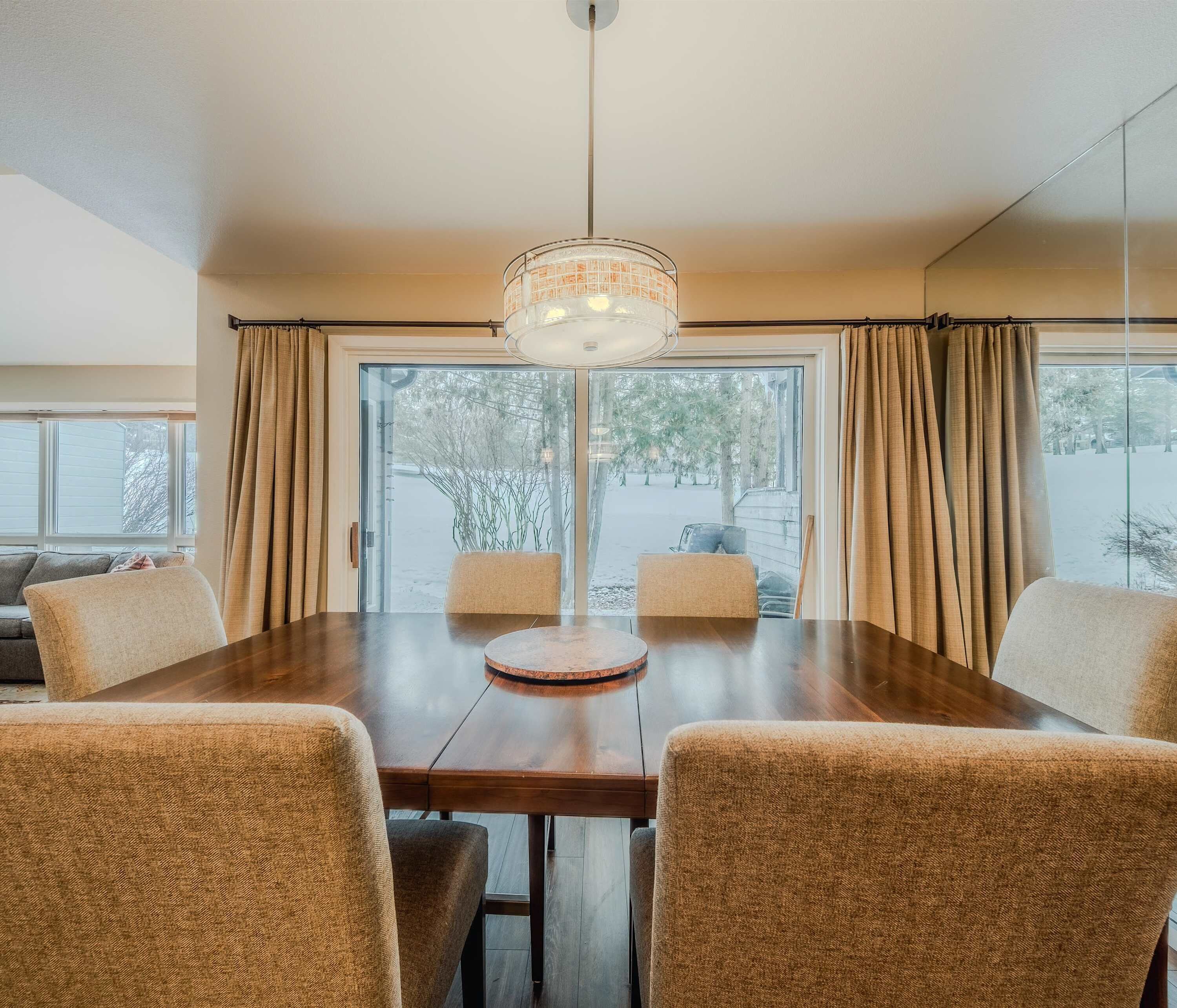 Un coin repas avec une table en bois et quatre chaises, un luminaire suspendu et de grandes fenêtres avec des rideaux.
