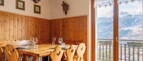 A wooden dining room with a large table and chairs, a wall-mounted clock, and a view of snow-capped mountains through a large window.