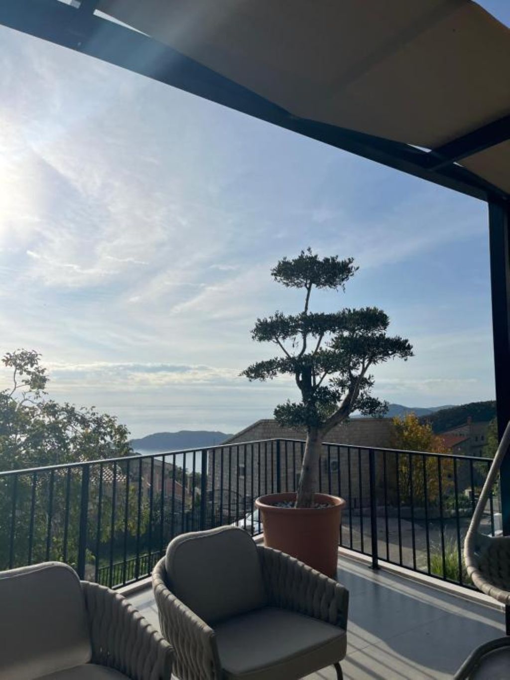 Un balcon agrémenté d’un bonsaï en pot, de chaises en osier et offrant une vue sur des collines lointaines.