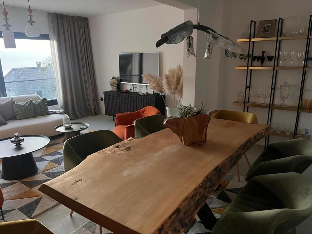 A modern living room with a wooden dining table, green and orange chairs, a sofa, a TV, and a bookshelf with glassware.