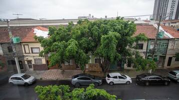 A street view with parked cars, a large tree, and buildings with red-tiled roofs.