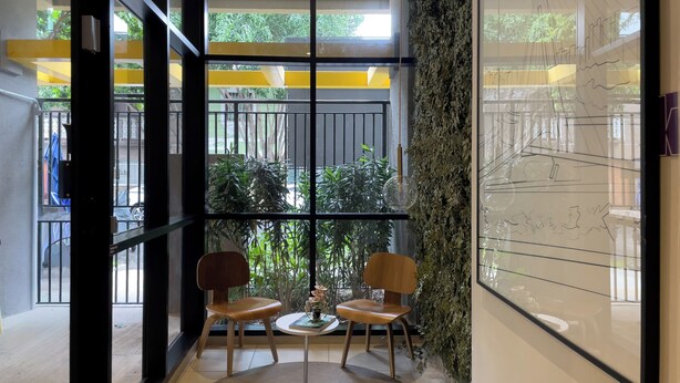 Un interior moderno con una pared de vidrio, dos sillas de madera, una mesita redonda y una pared verde con una lámpara colgante.
