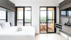 Una habitación de hotel moderna con cama, televisión, balcón con mesa y sillas, y ventanas grandes.