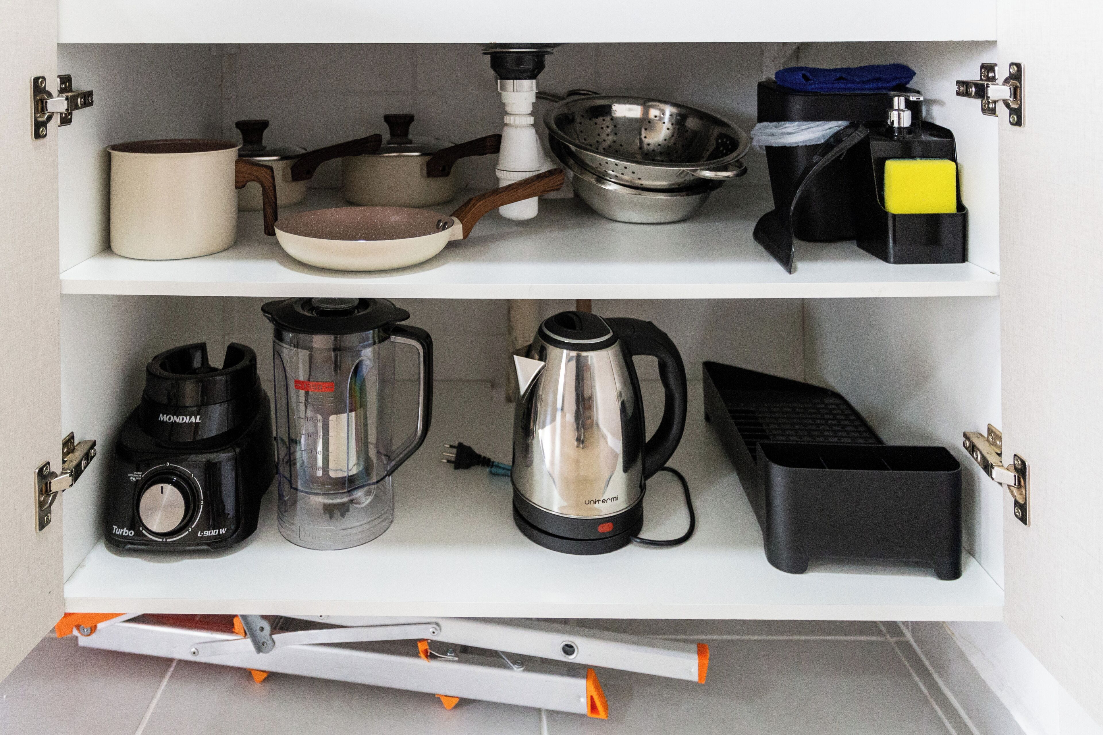 A kitchen with a white shelf containing a coffee maker, a kettle, a blender, a frying pan, a pot, a colander, and a cutting board.