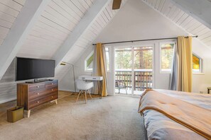Ein Schlafzimmer mit schrägverdeckter Decke, einem Bett, einem Schreibtisch, einem Fernseher und einem Balkon mit Vorhängen.