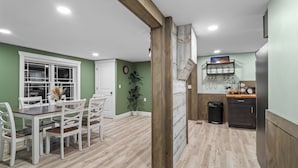A dining area with a wooden table and chairs, a kitchen with a refrigerator and shelves, and a clock on the wall.