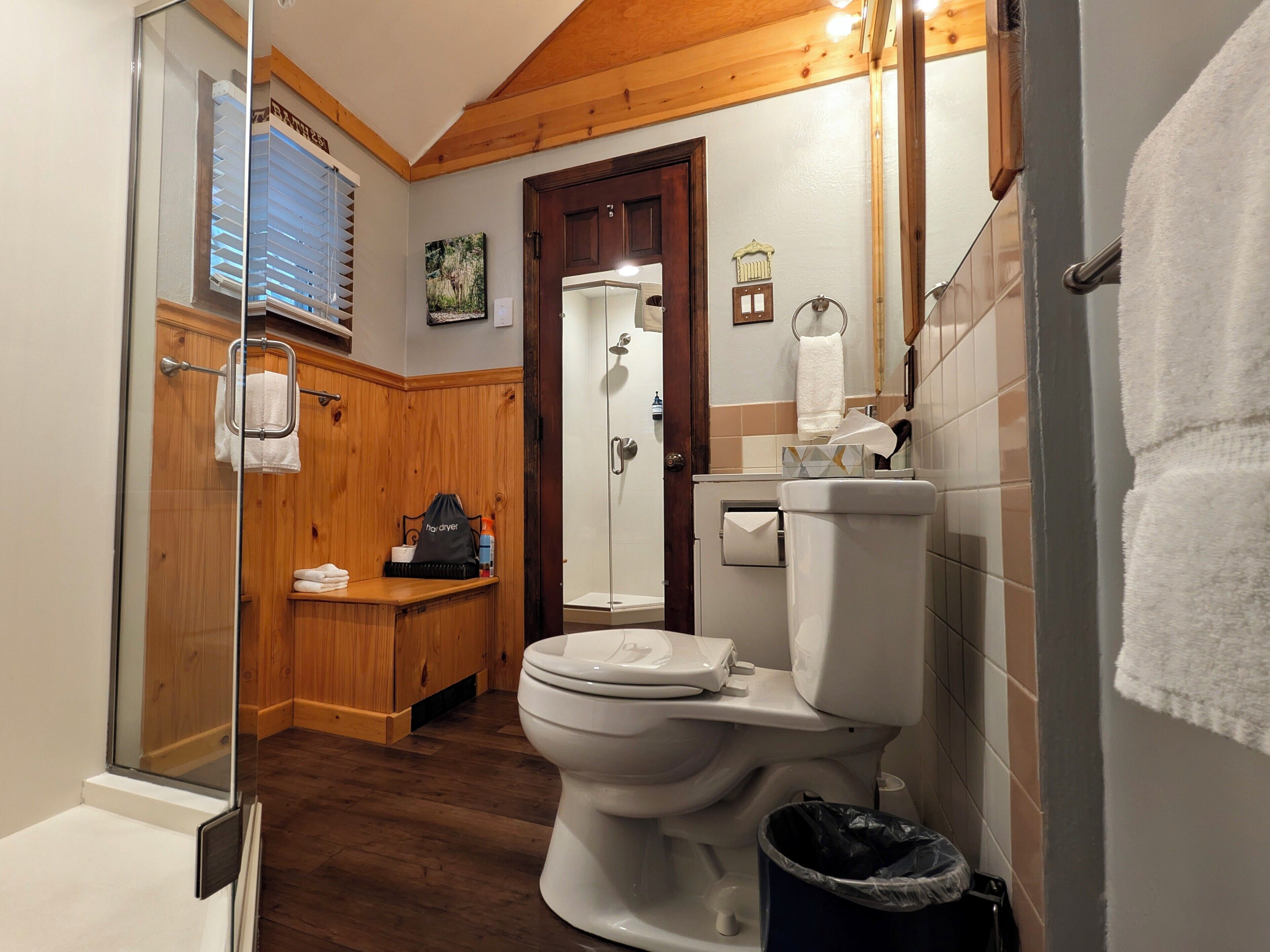Een badkamer met een houten vloer, toilet, douchecabine van glas en een bankje met handdoeken.
